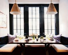 
                    
                        This kitchen dining nook is backdropped with black window panes, large bench seating, a black rectangle table, and two matching brass pendant lights.
                    
                